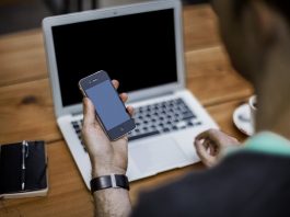 homem olha um celular em sua mão, com um laptop em sua frente numa mesa de madeira