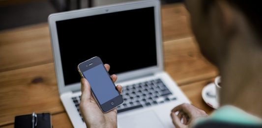 Telemarketing eleitoral homem olha um celular em sua mão, com um laptop em sua frente numa mesa de madeira