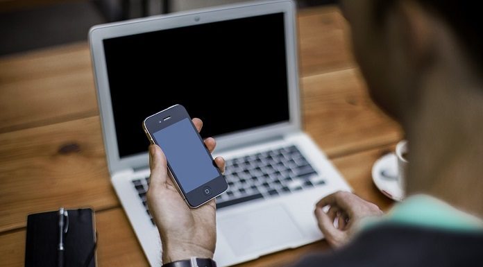 homem olha um celular em sua mão, com um laptop em sua frente numa mesa de madeira