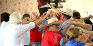 Idosos vestidos com trajes de arraiá fazem um túnel com as mãos em dança coletiva