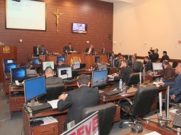 Salão da câmara de vereadores onde se vê ao fundo à esquerda a tribuna com um vereador falando e demais sentados em suas mesas com computadores de costas para a foto