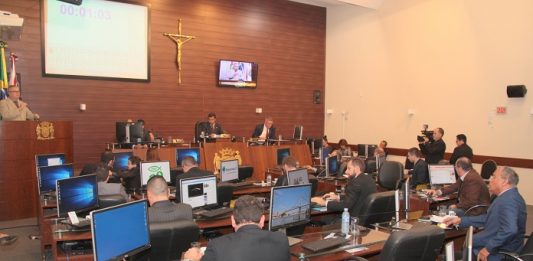 Salão da câmara de vereadores onde se vê ao fundo à esquerda a tribuna com um vereador falando e demais sentados em suas mesas com computadores de costas para a foto
