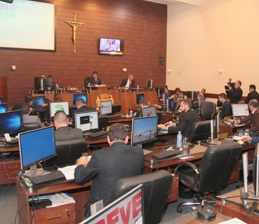 Salão da câmara de vereadores onde se vê ao fundo à esquerda a tribuna com um vereador falando e demais sentados em suas mesas com computadores de costas para a foto