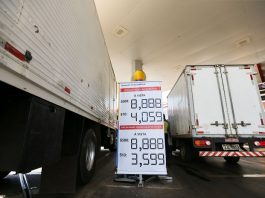 dois caminhões vistos pela parte de trás estacionados lado a lado em um posto com um banner mostrando os preços