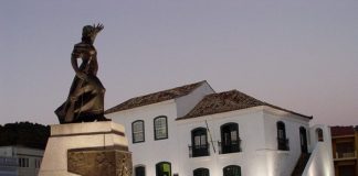 estátua de figura feminina iluminada em uma praça em frente a um casarão histórico