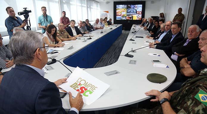 grande mesa de reunião em U com o governador sentado na ponta e visto de costas falando para duas dezenas de pessoas sentadas