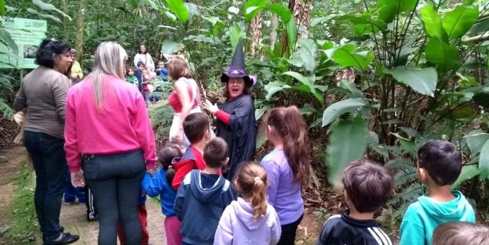 professora fantasiada de bruxa conversa com alunos em meio a uma mata