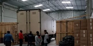 caminhão estacionado em galpão com caixas em volta e cinco pessoas paradas atrás