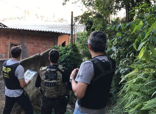 Três policiais vistos de costas usando coletes à prova de bala com escritos da polícia civil em frente à casas de favela