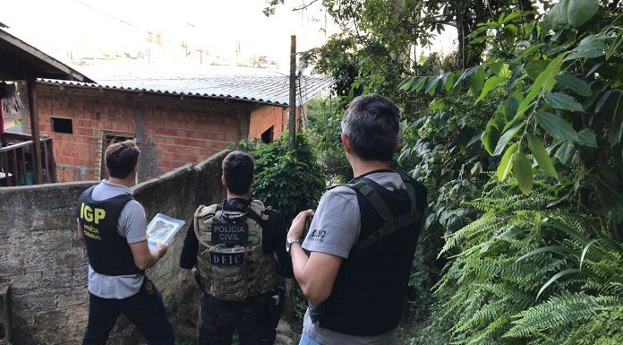 Três policiais vistos de costas usando coletes à prova de bala com escritos da polícia civil em frente à casas de favela