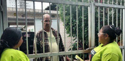 duas mulheres uniformizadas falam com cidadão através das grades de uma casa