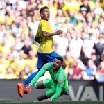 jogador firmino olha para fora da imagem e o goleiro adversário na mesma direção, caído no chão; grande torcida ao fundo