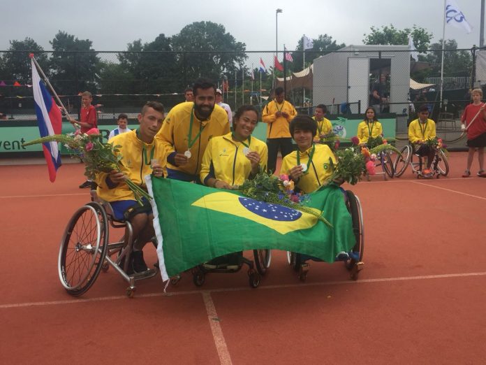 Três cadeirantes e o professor posam na quadra de tênis com medalhas e bandeira do Brasil