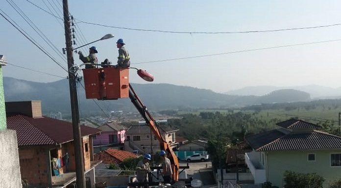 dois homem em uma grua, erguida a partir de um caminhão, mexem no alto do poste, em rua com várias casas