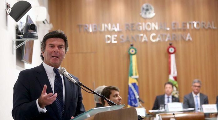 Ministro gesticula em uma bancada de madeira ao microfone com mesa ao fundo com pessoas sentadas e bandeira do Brasil e SC