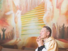 homem usando batina amarela e branca sentado em um banco de igreja olha sorridente para frente com as mãos em forma de oração perto do rosto