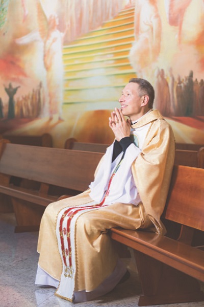 homem usando batina amarela e branca sentado em um banco de igreja olha sorridente para frente com as mãos em forma de oração perto do rosto