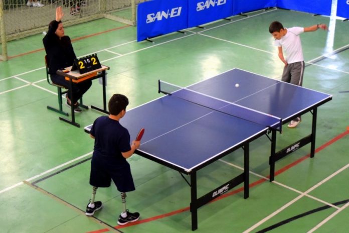 Para tenis Mesa de tênis vista de cima, onde jogam dois garotos, em que têm as pernas com próteses; ao lado uma juiza levanta a mão