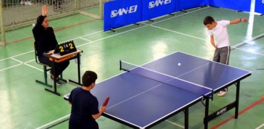 Florianópolis é sede dos Jogos Escolares Paradesportivos Mesa de tênis vista de cima, onde jogam dois garotos, em que têm as pernas com próteses; ao lado uma juiza levanta a mão