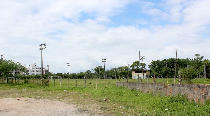foto de terreno baldio com campo de futebol e vegetação ao fundo, com alguns postes e um céu pouco nublado