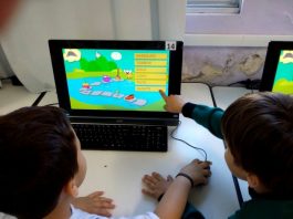 Duas crianças vistas de costas apontam para uma tela de computado onde há um jogo