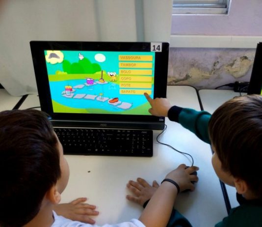 Duas crianças vistas de costas apontam para uma tela de computado onde há um jogo