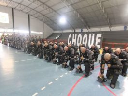 Repressão e presença do Estado em uma quadra de ginásio, tropa de homens carecas usando armaduras pretas se ajoelha em formação