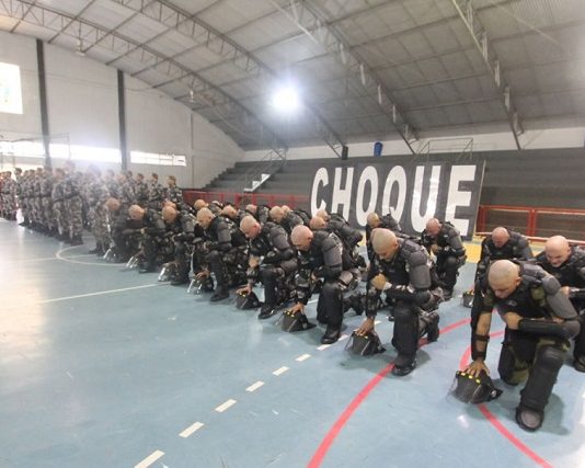 em uma quadra de ginásio, tropa de homens carecas usando armaduras pretas se ajoelha em formação