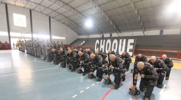 em uma quadra de ginásio, tropa de homens carecas usando armaduras pretas se ajoelha em formação