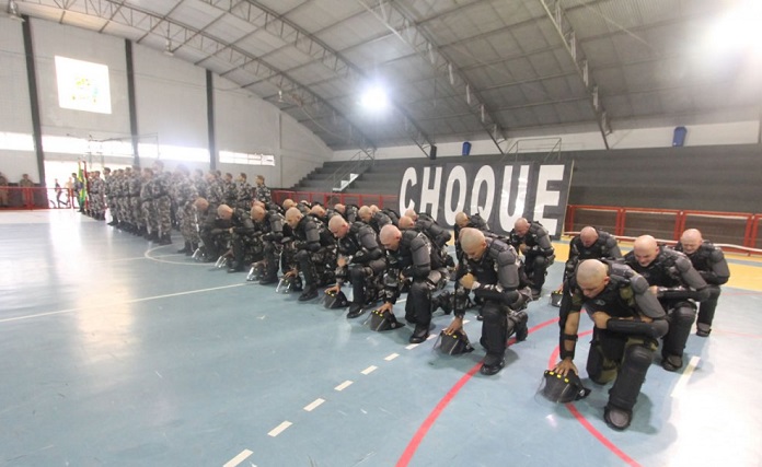 em uma quadra de ginásio, tropa de homens carecas usando armaduras pretas se ajoelha em formação