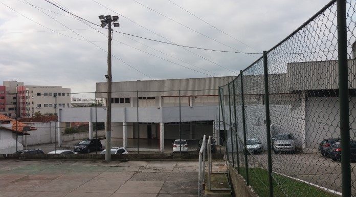 Vista geral do clube, com quadra de cimento em primeiro plano, cercada; atrás o prédio principal, com alguns carros estacionados na frente