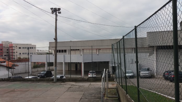 Vista geral do clube, com quadra de cimento em primeiro plano, cercada; atrás o prédio principal, com alguns carros estacionados na frente