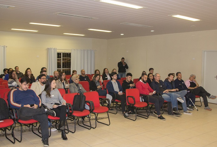 pessoas sentadas em cadeiras em uma sala grande, de noite, com janelas ao fundo