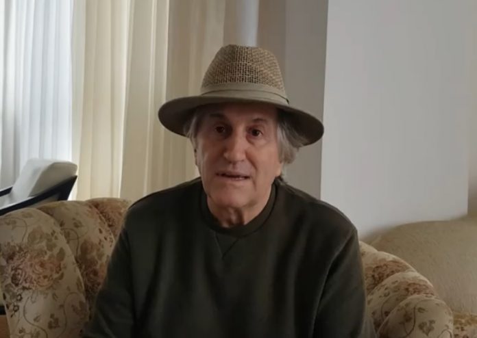 Pavan sentado em uma poltrona usa um chapéu e fala diretamente para a câmera