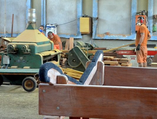 Violência e Saúde em pauta homens de uniforme laranja trabalhando em chão de fábrica, com diversos maquinários