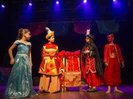 quatro crianças usando vestes caricatas de árabes olham entre si sobre um palco com baixa iluminação