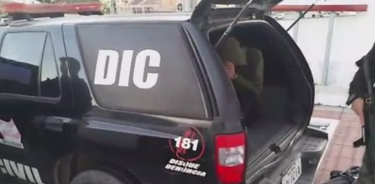 Operação na Maloca, em Florianópolis, cumpre 26 mandados de prisão Caminhonete preta da polícia onde se lê DIC no vidro, com a tampa do porta-malas aberta, onde há uma pessoa sentada e com capuz cobrindo o rosto; há dois policias ao lado da viatura