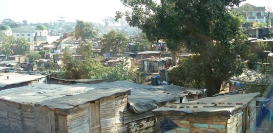 Desigualdade social contribui para o círculo vicioso da corrupção barracos de uma favela com algumas árvores; em primeiro plano, mulher negra se abaixa para lavar roupas
