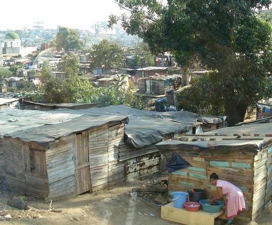 barracos de uma favela com algumas árvores; em primeiro plano, mulher negra se abaixa para lavar roupas