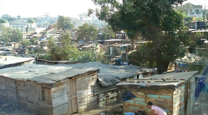 barracos de uma favela com algumas árvores; em primeiro plano, mulher negra se abaixa para lavar roupas