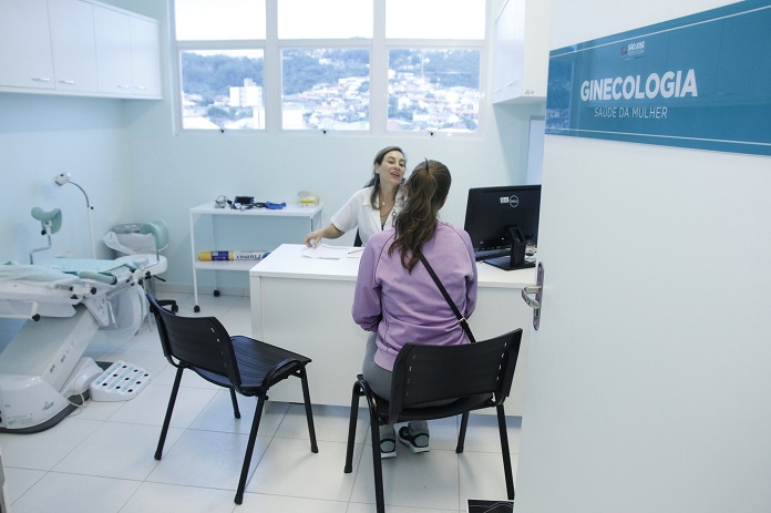 paciente mulher é vista de costas sentada em um consultório sendo atendida por uma médica; ao lado, na porta, está escrito 