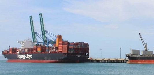 Empresas de SC estimam crescimento nas exportações em 2018 e 2019 navio cargueiro visto ao longe com containeres atracado em um porto em um dia de sol e mar calmo