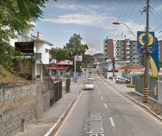 vista central da rua, com carros vindo ao fundo e comércios em volta num dia ensolarado