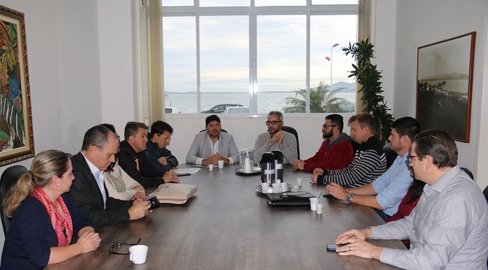 vista de central de uma mesa onde há pessoas sentadas em volta eu reunião; na ponta, o procurador; atrás dele, uma janela com vista para o mar