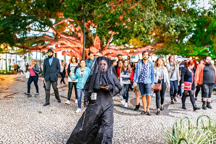 mulher trajada de uma típica viúva anda à frente de um grupo de pessoas na praça 15