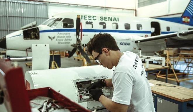 rapaz usando equipamento de segurança do trabalho mexe em peça de aviação com ferramentas em um hangar; há uma aeronave no fundo