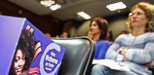cartilha da lei da maria da penha em primeiro plano sobre uma cadeira em auditório e mulheres sentadas ao fundo