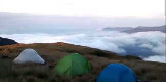 três barracas sobre uma relva grande e amarelada no alto de uma montanha com nuvens em volta