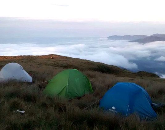 três barracas sobre uma relva grande e amarelada no alto de uma montanha com nuvens em volta