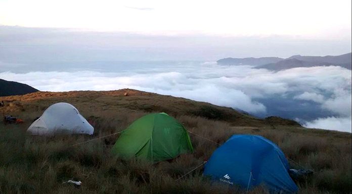 três barracas sobre uma relva grande e amarelada no alto de uma montanha com nuvens em volta